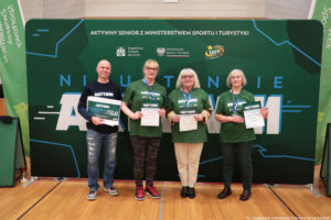 Grupa czterech dorosłych osób stojących w hali sportowej przed dużą zieloną ścianką eventową. Wszyscy stoją w jednym rzędzie i trzymają w dłoniach dyplomy/certyfikaty; osoba skrajnie z lewej trzyma dodatkowo duży bon. Tło to zielona ścianka z napisami i logotypami.