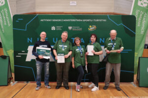 Grupa pięciu dorosłych osób stojących w hali sportowej przed dużą zieloną ścianką eventową. Wszyscy stoją w jednym rzędzie i trzymają w dłoniach dyplomy/certyfikaty; osoba skrajnie z lewej trzyma dodatkowo duży bon. Tło to zielona ścianka z napisami i logotypami.