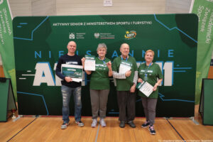 Grupa czterech dorosłych osób stojących w hali sportowej przed dużą zieloną ścianką eventową. Wszyscy stoją w jednym rzędzie i trzymają w dłoniach dyplomy/certyfikaty; osoba skrajnie z lewej trzyma dodatkowo duży bon. Tło to zielona ścianka z napisami i logotypami.