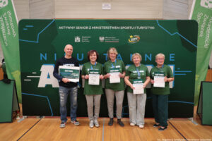 Grupa pięciu dorosłych osób stojących w hali sportowej przed dużą zieloną ścianką eventową. Wszyscy stoją w jednym rzędzie i trzymają w dłoniach dyplomy/certyfikaty; osoba skrajnie z lewej trzyma dodatkowo duży bon. Tło to zielona ścianka z napisami i logotypami.