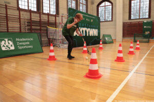 Aula sportowa. Seniorka w ciemnozielonej koszulce z białym napisem „Nieustannie aktywni” trzyma kij hokejowy i drybluje z piłką między biało-pomarańczowymi pachołkami.