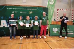 Grupa sześciu dorosłych osób stojących w hali sportowej przed dużą zieloną ścianką promocyjną. Cztery osoby na środku pozują do zdjęcia i trzymają dyplomy lub tablicę z nagrodą, a po prawej stronie mężczyzna w ciemnym stroju mówi do mikrofonu, trzymając kartki.