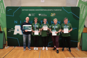 Grupa pięciu dorosłych osób stojących w hali sportowej przed dużą zieloną ścianką eventową. Wszyscy stoją w jednym rzędzie i trzymają w dłoniach dyplomy/certyfikaty; osoba skrajnie z lewej trzyma dodatkowo duży bon. Tło to zielona ścianka z napisami i logotypami.