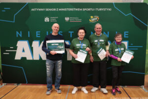 Grupa czterech dorosłych osób stojących w hali sportowej przed dużą zieloną ścianką eventową. Wszyscy stoją w jednym rzędzie i trzymają w dłoniach dyplomy/certyfikaty; osoba skrajnie z lewej trzyma dodatkowo duży bon. Tło to zielona ścianka z napisami i logotypami.