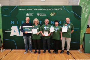 Grupa pięciu dorosłych osób stojących w hali sportowej przed dużą zieloną ścianką eventową. Wszyscy stoją w jednym rzędzie i trzymają w dłoniach dyplomy/certyfikaty; osoba skrajnie z lewej trzyma dodatkowo duży bon. Tło to zielona ścianka z napisami i logotypami.