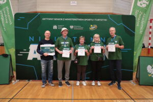 Grupa pięciu dorosłych osób stojących w hali sportowej przed dużą zieloną ścianką eventową. Wszyscy stoją w jednym rzędzie i trzymają w dłoniach dyplomy/certyfikaty; osoba skrajnie z lewej trzyma dodatkowo duży bon. Tło to zielona ścianka z napisami i logotypami.