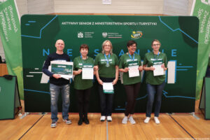 Grupa pięciu osób stojących na parkiecie hali sportowej na tle zielonej ścianki promocyjnej. Wszyscy uśmiechają się do zdjęcia. Cztery osoby (trzy kobiety po prawej i jedna kobieta druga od lewej) mają zielone koszulki z białym napisem „AKTYWNI” i medal na niebieskiej wstążce; trzymają w dłoniach kartki z napisem „CERTYFIKAT”. Osoba skrajnie po lewej (mężczyzna) ma ciemną bluzę i dżinsy i trzyma duży dokument z napisem „AKTYWNI VOUCHER 250 zł”. Na ściance w górnej części jest napis: „AKTYWNY SENIOR Z MINISTERSTWEM SPORTU I TURYSTYKI” oraz logotypy, w tym „Akademicki Związek Sportowy”, „Ministerstwo Sportu i Turystyki” i „Fundacja LOTTO”. Po bokach widać zielone pionowe banery z napisem „Akademicki Związek Sportowy”. W prawym dolnym rogu znajduje się podpis: „fot. Magdalena Seredyńska/Uniwersytet Szczeciński”.