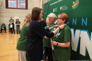 Ceremonia wręczania medali podczas wydarzenia sportowego w hali gimnastycznej. Na pierwszym planie młoda kobieta w czarnej sukience wiesza medal na szyi starszej kobiety w zielonej koszulce; starsza kobieta ma krótkie rude włosy, okulary i zegarek na lewej ręce. Obok stoją inni seniorzy w zielonych koszulkach, w tym starszy mężczyzna z białymi włosami i starsza kobieta z krótkimi siwymi włosami, uśmiechająca się. W tle widać parkiet hali, ścianę z oknami oraz kilka osób w zielonych koszulkach obserwujących uroczystość.
