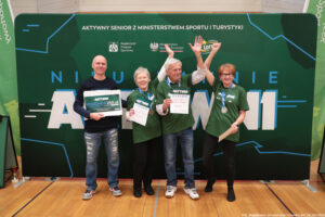 Grupa czterech osób stojących na sali gimnastycznej przed dużą zieloną ścianką promocyjną wydarzenia „Aktywny Senior”. Na środku stoją trzy starsze osoby w zielonych koszulkach i z medalami na szyi; dwie z nich trzymają dyplomy, a mężczyzna pośrodku ma uniesione ręce w górę. Po lewej stoi łysy mężczyzna w czarnej bluzie i dżinsach, trzyma tabliczkę z napisem „VOUCHER 250 zł”. Na górze ścianki widoczny jest napis: „AKTYWNY SENIOR Z MINISTERSTWEM SPORTU I TURYSTYKI” oraz kilka logotypów, m.in. „Akademicki Związek Sportowy” i „Ministerstwo Sportu i Turystyki”. W tle, dużymi literami, widać słowo „AKTYWNI”. W prawym dolnym rogu zdjęcia jest podpis: „fot. Magdalena Seredyńska/Uniwersytet Szczeciński”.