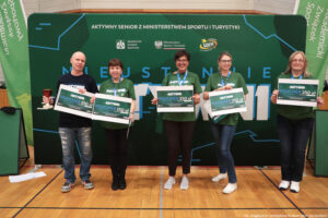 Grupa pięciu osób stojących w hali sportowej na tle dużej zielonej ścianki promocyjnej. Na ściance u góry widoczny napis: „AKTYWNY SENIOR Z MINISTERSTWEM SPORTU I TURYSTYKI” oraz logotypy (m.in. Akademicki Związek Sportowy, Ministerstwo Sportu i Turystyki, Fundacja LOTTO). Każda z osób trzyma duży, biało-zielony czek/voucher z napisem „AKTYWNI VOUCHER 250 zł”. Dodatkowo osoba skrajnie z lewej trzyma w drugiej dłoni niewielki puchar. Cztery osoby mają zielone koszulki z napisem „AKTYWNI” i identyfikatory na smyczach, jedna (z lewej) ma czarną koszulkę. Podłoga to drewniany parkiet z liniami boiskowymi. W prawym dolnym rogu znajduje się podpis: „fot. Magdalena Seredyńska / Uniwersytet Szczeciński”.