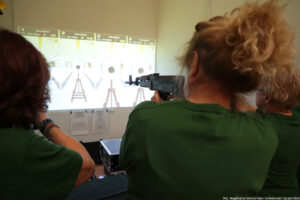 Sylwetki trzech osób tyłem. Osoby trzymają broń laserową i celują do punktów na ścianie, które wyznacza rzutnik.