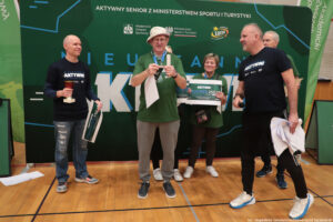 Grupa pięciu osób na hali sportowej na tle dużej zielonej ścianki promocyjnej z napisem „AKTYWNY SENIOR Z MINISTERSTWEM SPORTU I TURYSTYKI” oraz logotypami (m.in. Akademicki Związek Sportowy, Ministerstwo Sportu i Turystyki, Fundacja LOTTO). W centrum stoi starszy mężczyzna w jasnym kapeluszu, zielonej koszulce i szarych spodniach dresowych; na szyi ma medal i trzyma w rękach papier lub wstęgę. Po lewej stoi mężczyzna w czarnej koszulce z napisem „AKTYWNI”, trzyma puchar i prostokątny plakat/tablę. Po prawej stoi mężczyzna w czarnej koszulce „AKTYWNI”, z mikrofonem w jednej dłoni i plikiem kartek w drugiej. Z tyłu, nieco po prawej, stoi kobieta w zielonej koszulce, trzyma duży bon/plakat z widocznym napisem „DECATHLON VOUCHER 250”. Z tyłu po prawej częściowo widać jeszcze jednego starszego mężczyznę. Podłoże to drewniana podłoga boiska; w prawym dolnym rogu jest mały napis: „fot. Magdalena Seredyńska/Uniwersytet Szczeciński”.