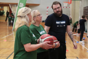 Aula sportowa. Dwie seniorki (ujęcie bokiem) w ciemnozielonych koszulkach z białym napisem „Nieustannie aktywni” trzymają piłki do kosza i rozmawiają z jurorem (przodem do kadru). W tle inni uczestnicy igrzysk seniora.