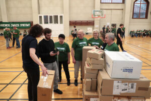 Grupa ludzi stojąca na hali sportowej obok stosu kartonowych pudeł. Na pierwszym planie po prawej stronie ułożone są większe i mniejsze paczki, w tym jedno duże białe pudło z nadrukiem. Obok paczek stoi kilka osób: starsi i młodsi dorośli, część w zielonych koszulkach z napisem „AKTYWNI”, jedna osoba w ciemnej koszulce opiera dłoń na dużym kartonie, a mężczyzna w czarnej bluzie trzyma telefon i coś sprawdza. W tle widać parkiet z liniami do gier, kosz do koszykówki, ławki oraz więcej osób w podobnych zielonych koszulkach; po lewej stronie jest zielony baner z napisem „AKADEMICKI ZWIĄZEK SPORTOWY”. W prawym dolnym rogu widnieje podpis: „fot. Magdalena Seredyńska/Uniwersytet Szczeciński”.