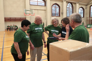 Grupa starszych osób w zielonych koszulkach sportowych rozmawia w sali gimnastycznej. Na koszulkach widoczny jest duży napis „AKTYWNI” (nad nim mniejszy tekst, nieczytelny). W centrum stoi łysy mężczyzna w zielonej koszulce i jasnych spodniach, obok niego kobieta z jasnymi, kręconymi włosami i okularami, też w zielonej koszulce. Po lewej jest niższa kobieta z krótkimi, ciemnymi włosami w zielonej koszulce. Po prawej stoi mężczyzna z siwymi włosami w zielonej koszulce, odwrócony bokiem. Między nimi jest kobieta w ciemnej koszulce, trzyma długopis i coś notuje na podkładce/klipbordzie. W prawym dolnym rogu kadru znajduje się duże kartonowe pudełko zaklejone taśmą. Tło to hala sportowa z drewnianą podłogą, liniami boiska i koszem do koszykówki; dalej widać siedzące osoby w podobnych zielonych koszulkach. W prawym dolnym rogu jest podpis: „fot. Magdalena Seredyńska/Uniwersytet Szczeciński”.
