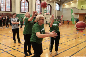 Aula sportowa. Trzy seniorki (ujęcie bokiem) w ciemnozielonych koszulkach z białym napisem „Nieustannie aktywni” rzucają przed siebie piłkami do kosza. W tle inni uczestnicy igrzysk seniora.