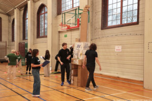 Sala gimnastyczna z parkietem i koszem do koszykówki oraz wysoką wieżą z kartonów ustawioną pod tablicą kosza. Na wieży widać różne pudełka i długie, wąskie kartony ustawione pionowo, a na samej górze kilka mniejszych kartonów. Obok stoją i przechodzą osoby w sportowych ubraniach; jedna osoba trzyma clipboard z kartkami, a inna podnosi rękę w stronę konstrukcji. W tle są duże okna z widokiem na drzewa oraz siatka ochronna na ścianie. Na ścianie po prawej jest kartka z napisem: „7 Kartony”. W prawym dolnym rogu widoczny podpis: „fot. Magdalena Seredyńska / Uniwersytet Szczeciński”.