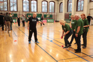 Aula sportowa. Po prawej czworo seniorów (3 kobiety, 1 mężczyzna – ujęcie bokiem) w ciemnozielonych koszulkach z białym napisem „Nieustannie aktywni” rzucają małymi czerwonymi piłeczkami. Po lewej juror. W tle inni uczestnicy igrzysk seniora.