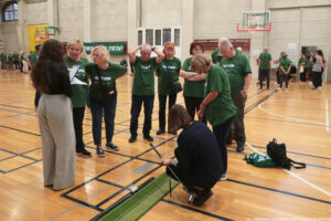 Aula sportowa. Ośmioro seniorów (5 kobiet, 3 mężczyzn) w ciemnozielonych koszulkach z białym napisem „Nieustannie aktywni” grają w minigolfa. Tyłem do kadru dwie jurorki. W tle inni uczestnicy igrzysk seniora.