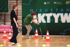 Aula sportowa. Seniorka w ciemnozielonej koszulce trzyma kij hokejowy i drybluje z piłką między biało-pomarańczowymi pachołkami. Po lewej jurorka (tyłem do kadru). W tle ciemnozielona ścianka z napisem „Nieustannie aktywni”.