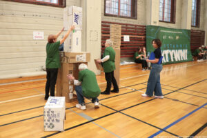 Aula sportowa. Troje seniorów (1 mężczyzna i 2 kobiety) w ciemnozielonych koszulkach zbuduje wieżę z kartonów podczas igrzysk seniorów. Po prawej jurorka.