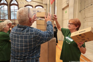Aula sportowa. Dwoje seniorów (1 mężczyzna – tyłem do kadru i 1 kobieta w ciemnozielonej koszulce z białym napisem „Nieustannie aktywni”) buduje wieżę z kartonów podczas igrzysk seniorów. Po lewej w tle dwoje innych uczestników wydarzenia.