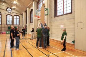 Aula sportowa. Czworo seniorów (1 mężczyzna i 3 kobiety) w ciemnozielonych koszulkach z budują podczas igrzysk seniorów wysoką wieżę z kartonów. Po lewej dwoje jurorów.