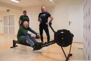 Senior siedzący na ergometrze wioślarskim wykonuje zadanie podczas igrzysk seniorów. Za nim stoi drugi senior (obaj ubranie w ciemnozielone koszulki z białym napisem „Nieustannie aktywni”), obok juror z teczką.