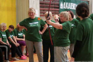 Aula sportowa. Cztery seniorki w ciemnozielonych koszulkach z białym napisem „Nieustannie aktywni” – jedna trzyma łuk i celuje do punktu. W tle inne uczestniczki igrzysk seniorów.
