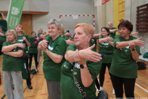 Aula sportowa. Kilka seniorek w ciemnozielonych koszulkach z białym napisem „Nieustannie aktywni” stoi w pozycji, w której jedna ręka podtrzymuje drugą – skierowaną w przeciwną stronę. W tle inni uczestnicy igrzysk seniorów.