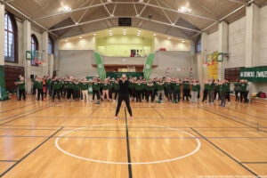 Aula sportowa. Ponad 50-osobowa grupa seniorów w ciemnozielonych koszulkach stoi w pozycji, w której jedna ręka podtrzymuje drugą – skierowaną w przeciwną stronę. Na pierwszym planie tyłem do kadru trenerka prezentująca ćwiczenia podczas igrzysk seniorów.