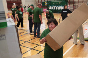 Grupa osób na hali sportowej podczas zajęć lub wydarzenia ruchowego. W centrum na pierwszym planie starsza kobieta o jasnej karnacji, w zielonej koszulce, niesie duże prostokątne kartonowe pudełko ustawione po skosie. Na podłodze hali widać linie boiska oraz rozstawione małe pachołki i znaczniki. W tle stoi kilka osób, głównie w zielonych koszulkach z białym napisem, część rozmawia lub czeka; jedna osoba ma zieloną czapkę. Po prawej stronie stoi kobieta w czarnej koszulce z białym napisem, trzyma długi kij lub laskę. Z tyłu znajduje się duży zielony baner z widocznym napisem „AKTYWN…” (reszta zasłonięta) oraz logo LOT. W prawym dolnym rogu jest podpis: „fot. Magdalena Seredyńska/Uniwersytet Szczeciński”.