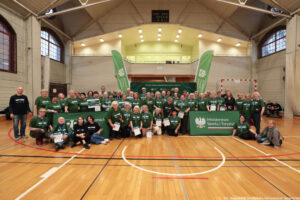 Aula sportowa. Ponad 50-osobowa grupa seniorów w ciemnozielonych koszulkach pozuje do zdjęcia z dyplomami igrzysk seniorów.