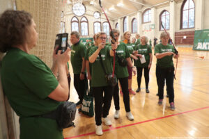 Grupa starszych osób w zielonych koszulkach podczas zajęć z łucznictwa w hali sportowej. W centrum kobieta w okularach trzyma łuk i celuje przed siebie; strzała ma pomarańczową przyssawkę na końcu. Po prawej inna kobieta także trzyma łuk z taką samą strzałą. Po lewej stronie kadru widać kobietę fotografującą telefonem. W tle są wysokie, łukowate okna, kosz do koszykówki i baner z dużym napisem „AKTYWNI”. Na dole zdjęcia jest drobny podpis: „fot. Magdalena Seredyńska / Uniwersytet Szczeciński”.