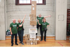 Aula sportowa. Czworo seniorów (1 mężczyzna i 3 kobiety) w ciemnozielonych koszulkach z białym napisem „Nieustannie aktywni” prezentuje zbudowaną podczas igrzysk seniorów wieżę z kartonów.