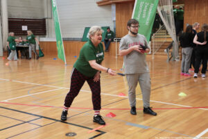 Hala sportowa z drewnianą podłogą i grupą osób biorących udział w zajęciach lub wydarzeniu sportowym. Na pierwszym planie starsza kobieta o jasnej cerze i krótkich siwych włosach, w zielonej koszulce i ciemnych spodniach we wzór, jest w pochylonej pozycji i wykonuje ruch ręką jak przy rzucie. Ma czarne buty i skarpetki w biało-czerwone paski. Obok stoi młody mężczyzna o jasnej cerze, w okularach, z krótkimi ciemnymi włosami i zarostem, ubrany w szary T-shirt z napisem „ESPAÑA” i szare spodnie, trzyma w rękach kilka małych przedmiotów. Na podłodze rozstawione są kolorowe pachołki treningowe (czerwone, żółte, niebieskie). W tle widać inne osoby w zielonych koszulkach przy stoliku oraz grupę osób po prawej stronie, a także zielone pionowe banery z białym napisem „Akademicki Związek Sportowy”. W prawym dolnym rogu jest podpis: „fot. Magdalena Seredyńska / Uniwersytet Szczeciński”.