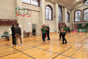 Sala gimnastyczna z boiskiem do koszykówki i grupą ludzi podczas zajęć sportowych. W kadrze widać kosz do koszykówki po lewej stronie; piłka jest w powietrzu tuż nad obręczą. Na parkiecie stoi kilka osób dorosłych, część w zielonych koszulkach, część w ciemnych ubraniach. Dwie osoby po prawej trzymają piłki do koszykówki. W tle ustawione są pachołki treningowe, a przy ścianie wiszą drabinki gimnastyczne. Po prawej stronie widać zielone banery z napisami „Fundacja LOTTO” oraz „Akademicki Związek Sportowy”. Na ścianie są wysokie, łukowate okna. Na dole zdjęcia jest podpis: „fot. Magdalena Seredyńska / Uniwersytet Szczeciński”.