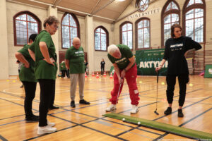 Grupa osób w sali gimnastycznej podczas zajęć sportowych z elementami gry w mini-golfa. Widać kilka osób starszych w zielonych koszulkach z nadrukiem oraz młodą kobietę w czarnym stroju; wszyscy stoją wokół wąskiego zielonego toru na podłodze. Starszy mężczyzna z siwymi włosami, w czerwonych spodniach i białych butach, pochyla się i uderza kijem golfowym w kierunku białych piłek leżących na torze. Pozostali uczestnicy obserwują, część ma ręce na biodrach. W tle są pachołki i małe znaczniki na parkiecie, duży zielony baner z napisem „AKTYWNI”, a wysoko nad oknami widoczny jest napis „UNIWERSYTET”. Na dole zdjęcia znajduje się podpis: „fot. Magdalena Seredyńska/Uniwersytet Szczeciński”.