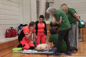 Grupa osób w sali gimnastycznej ćwicząca pierwszą pomoc na fantomie do resuscytacji leżącym na macie. Kilka osób w zielonych koszulkach klęczy i pochyla się nad fantomem, a dwoje ratowników w pomarańczowo-czarnych uniformach siedzi i stoi obok, obserwując i instruując. Na podłodze, przy macie, leżą rozłożone akcesoria medyczne i opakowania, a obok stoi zielone etui (prawdopodobnie na sprzęt typu AED) oraz czerwona torba ratownicza. W tle widać drewniane ławki przy ścianie i siatkę sportową. W prawym dolnym rogu jest podpis: „fot. Magdalena Seredyńska/Uniwersytet Szczeciński”.”