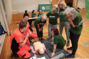 Grupa osób w hali sportowej podczas szkolenia z pierwszej pomocy przy fantomie do resuscytacji leżącym na macie. W centrum na podłodze znajduje się manekin–tułów w kolorze jasnej skóry z głową. Obok klęczy ratownik w pomarańczowo-czarnym uniformie i pokazuje coś gestami, a przy nim kobieta w podobnym pomarańczowym stroju. Wokół fantomu stoi i kuca kilka kobiet w zielonych koszulkach z napisem „AKTYWNI”, uważnie słuchając i obserwując; jedna ma kręcone siwe włosy i okulary, inne mają krótkie lub dłuższe włosy. W tle widać stolik z kilkoma osobami siedzącymi oraz zielony baner z napisem „AZS Akademicki Związek Sportowy”. Na dole zdjęcia widoczny jest podpis: „fot. Magdalena Seredyńska/Uniwersytet Szczeciński”.