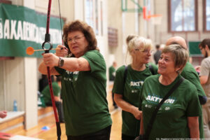 Grupa osób w zielonych koszulkach podczas zajęć sportowych w hali gimnastycznej. Kobieta na pierwszym planie trzyma łuk i celuje, mając naciągniętą cięciwę; na końcu strzały widoczna jest pomarańczowa przyssawka. Ma kręcone, brązowe włosy, okulary i na lewym nadgarstku opaskę sportową. Obok stoją dwie starsze kobiety w podobnych koszulkach; jedna ma jasne blond włosy związane w kucyk i okulary, druga ma krótkie siwe włosy i uśmiecha się. W tle widać rozmytych uczestników, fragment kosza do koszykówki oraz zielony baner na ścianie z dużymi białymi literami. Na dole po prawej jest mały podpis: „fot. Magdalena Seredyńska/Uniwersytet Szczeciński”.