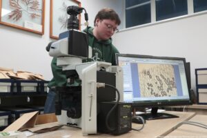 Noc Biologów 2026. Ujęcie bokiem. Na pierwszym planie mikroskop, za nim ekran monitora z widokiem spod mikroskopu. W tle prowadzący warsztaty w zielonej bluzie z białym logiem Uniwersytetu Szczecińskiego.