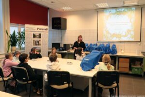 Noc Biologów 2026. Duży biały stół, dookoła którego siedzą dzieci – w większości tyłem do kadru. W tle pośrodku prowadząca, po lewej biały roll up na temat zagrożenia dla wód i po prawej na ścianie ekran z prezentacją.