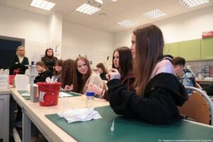 Noc Biologów 2026. Sala laboratoryjna. Ujęcie bokiem. Uczniowie siedzą za dwoma długimi stołami ustawionymi w rzędach. Na pierwszym planie dziewczyna zadająca pytanie (bokiem do kadru). W tle prowadzące warsztaty.