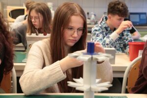 Noc Biologów 2026. Sala laboratoryjna. Ujęcie przodem. Uczniowie siedzą za stołami ustawionymi w rzędach. Na pierwszym planie dziewczyna przyglądająca się preparatowi.