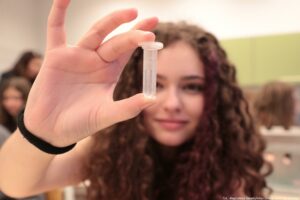 Noc Biologów 2026. Na pierwszym planie dłoń prezentująca probówkę. W tle trzymająca ją dziewczyna w ciemnych kręconych włosach.