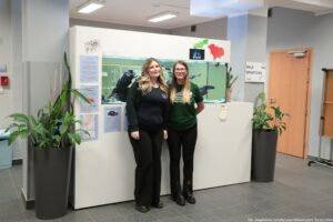 Noc Biologów 2026. Dwie studentki w koszulkach Uniwersytetu Szczecińskiego pozują do zdjęcia. W tle akwarium z rybami.