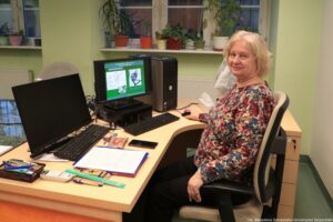 Noc Biologów 2026. Ujęcie bokiem. Kobieta siedząca za biurkiem. Na którym stoją dwa monitory (tylko jeden jest włączony) z dwiema klawiaturami.