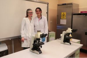 Noc Biologów 2026. Sala laboratoryjna. Ujęcie bokiem. Dwie laborantki w białych fartuchach pozują do zdjęcia. Przed nimi na białym stole stoją dwa mikroskopy.