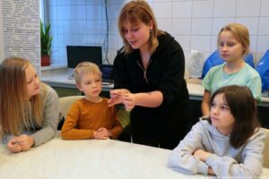 Noc Biologów 2026. Duży biały stół, dookoła którego siedzi czworo dzieci zaciekawionych tym, co pokazuje im prowadząca (w środku).